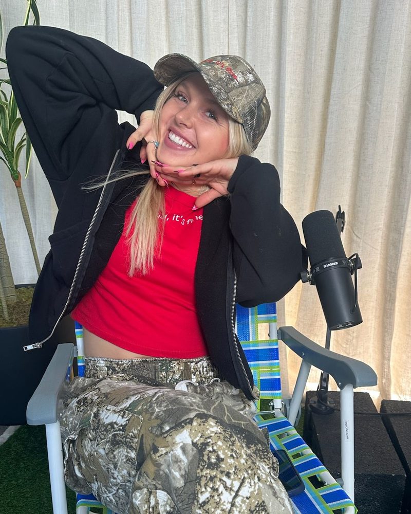 Ash grinning in a camo cap and red tee, leaning on her hands in a podcast studio chair